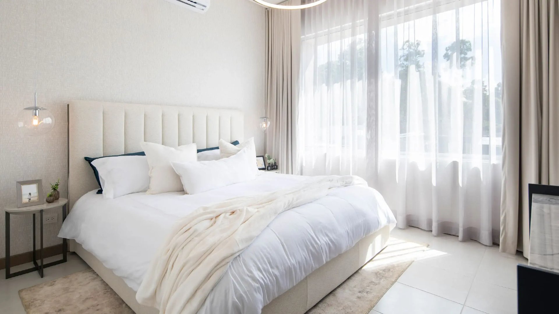 Render del dormitorio principal. Muestra una amplia cama con cabecera capitoneada beige, ropa de cama blanca, modernas lámparas colgantes de vidrio y un gran ventanal de piso a techo con cortinas que permiten el paso de la luz natural.