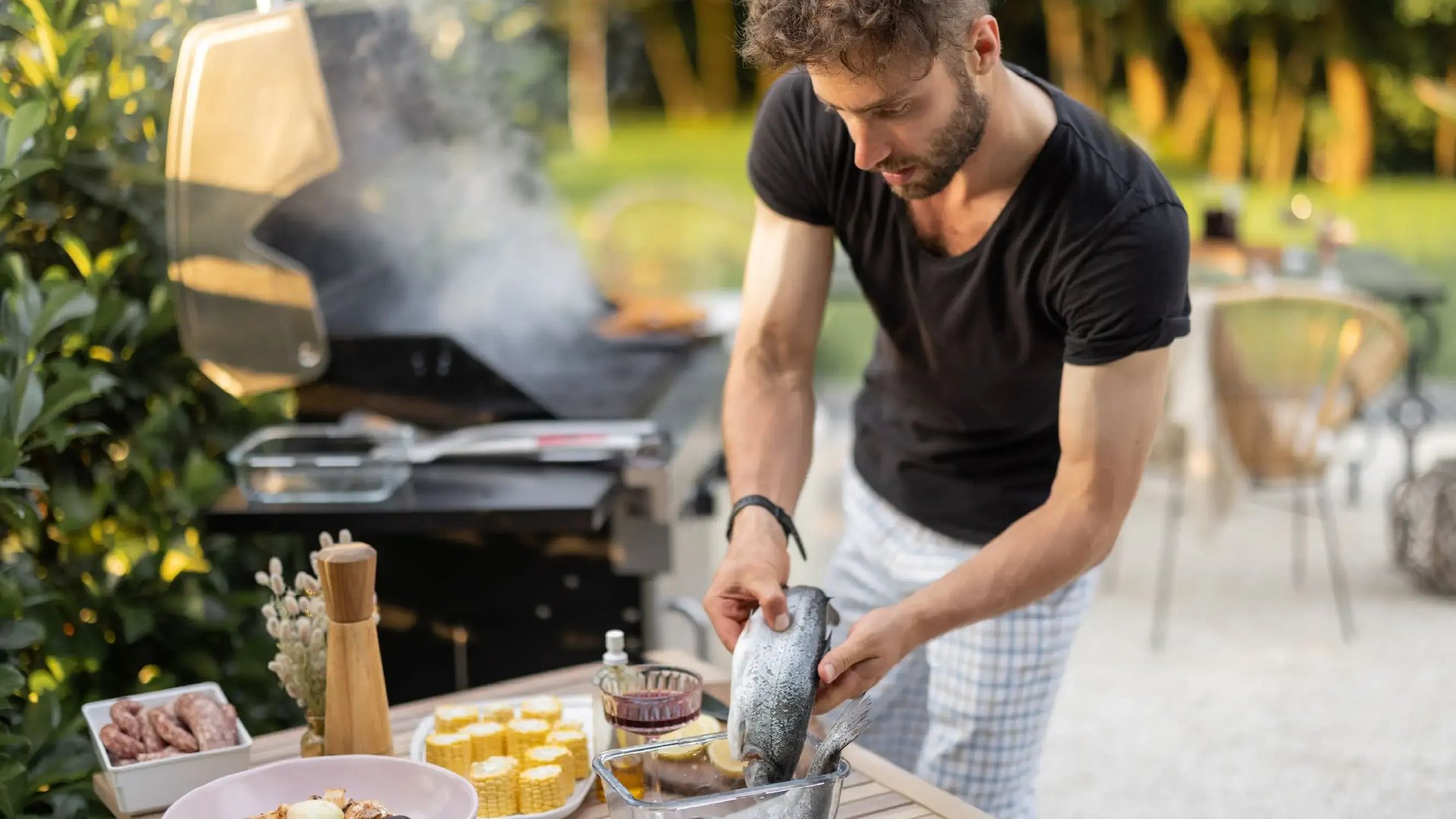 Hombre joven preparando comida en una parrilla de acero inoxidable en un día soleado, con una mesa servida con maíz, brochetas y vino al fondo.