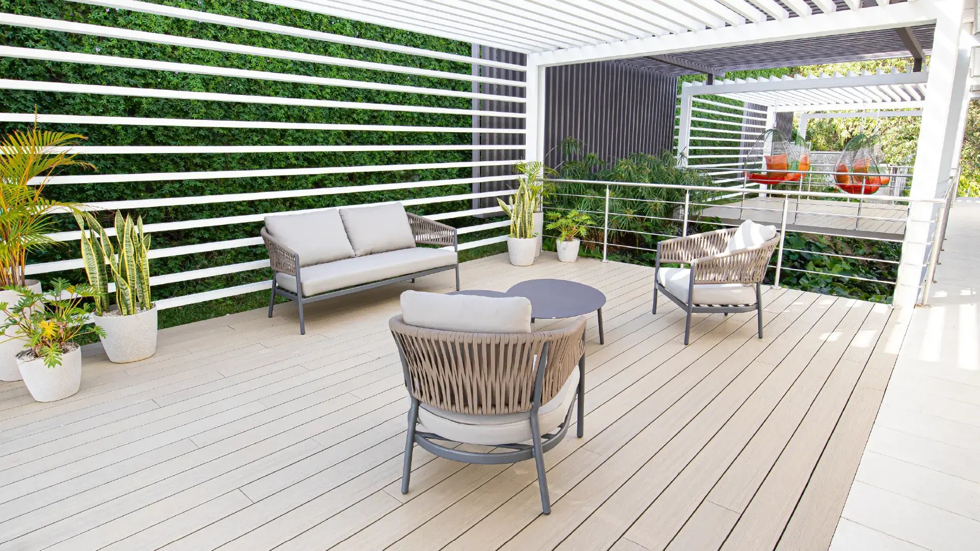 Zona de descanso al aire libre en la Casa Club de Condominio Naz, Alajuela. Muestra mobiliario de terraza moderno con cojines grises, mesa de centro, jardineras con plantas y una estructura de pérgola blanca techada.