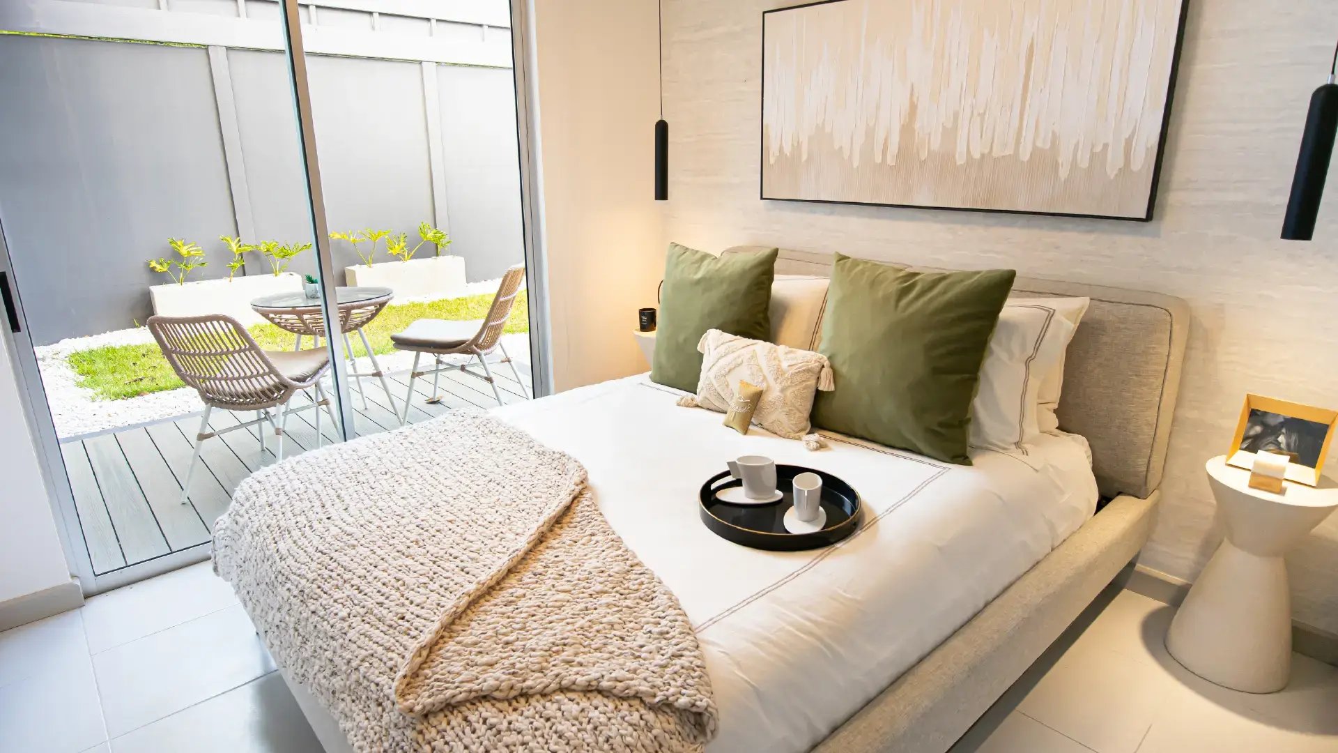 Dormitorio principal moderno en Condominio Naz, Alajuela. Muestra cama queen con edredón blanco y manta tejida beige, dos almohadas verdes, pared texturizada gris, lámparas colgantes negras y salida directa al jardín privado mediante puerta de vidrio.