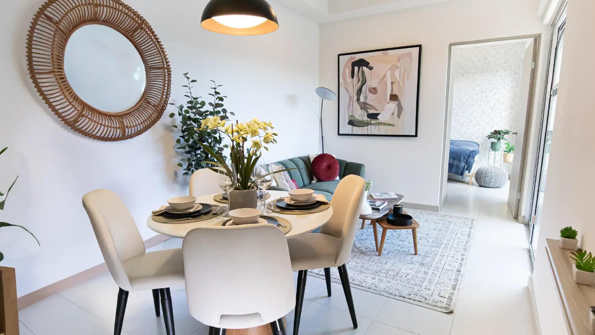 Área de comedor moderna en casa modelo de Condominio Naz, Alajuela. Incluye mesa circular blanca, sillas tapizadas beige, espejo de ratán y vista integrada hacia la sala de estar y el patio.