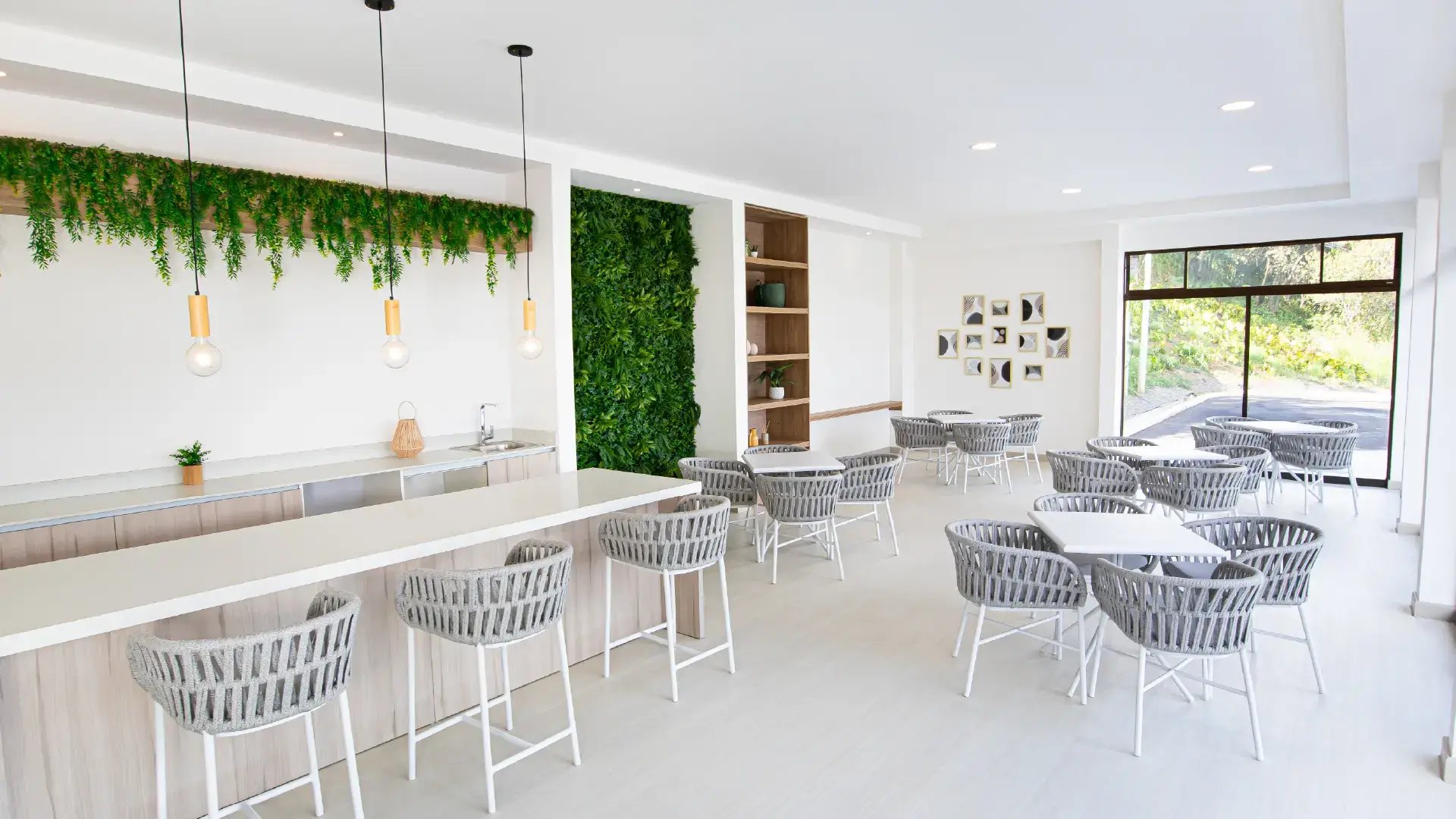 Área de bar y eventos sociales en Condominio Naz, Alajuela. Muestra una barra moderna con sobres blancos, bancos altos de tejido gris, jardín vertical en la pared y mesas de café en un ambiente iluminado y elegante.
