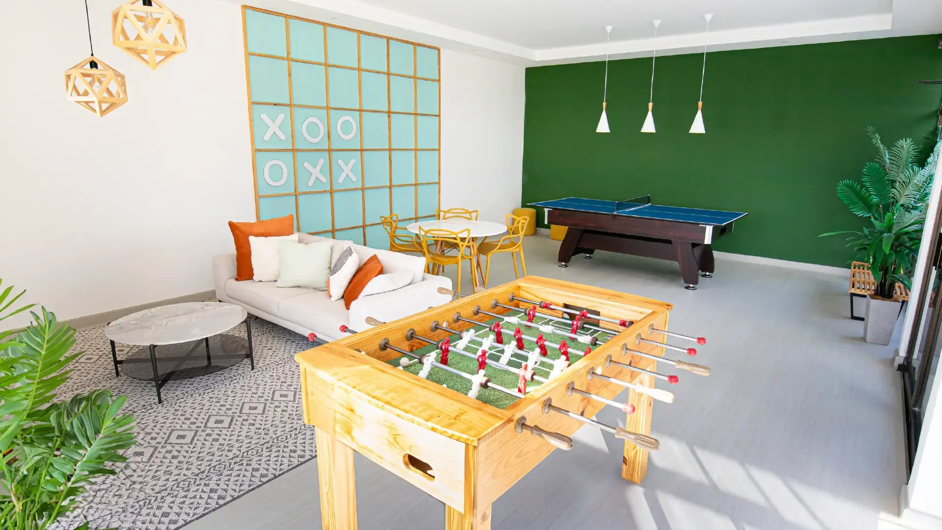Sala de juegos equipada en Condominio Naz, Alajuela. Muestra mesa de futbolín de madera, mesa de ping pong, zona de estar con sofá y un juego de 