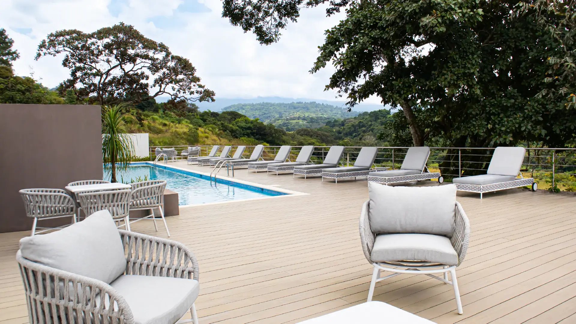 Deck de madera en la zona de piscina de Condominio Naz, Alajuela. Muestra mobiliario de exterior de lujo con sillones tejidos, camastros alineados frente a la piscina y una vista panorámica hacia las montañas de Alajuela.
