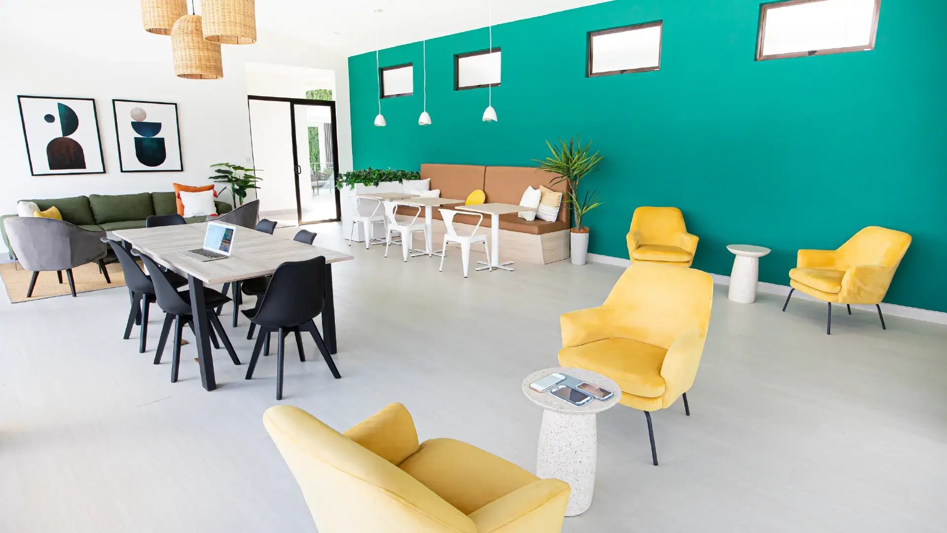 Área de coworking y sala social moderna en Condominio Naz, Alajuela. Muestra mesa de trabajo con laptop, sillas de diseño, cómodos sillones amarillos y decoración contemporánea con una pared turquesa vibrante.