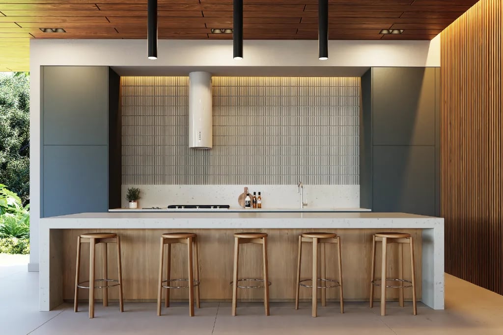 Render de cocina social moderna en casa club. Destaca una isla con sobre de cuarzo blanco, cinco bancos de madera, gabinetes minimalistas en gris oscuro, salpicadero con textura vertical e iluminación LED cálida en el cielorraso.