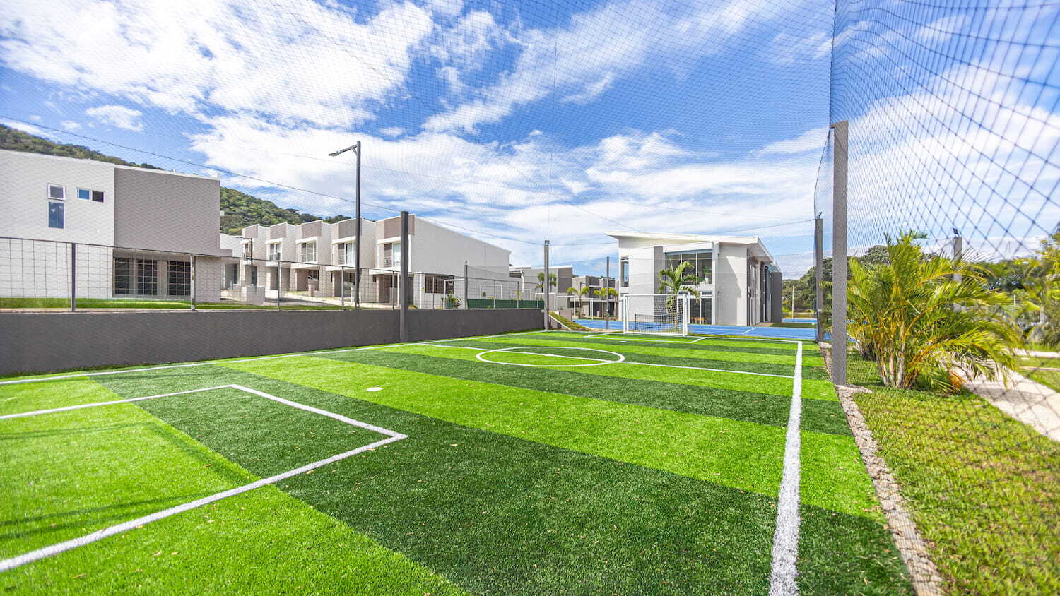 Cancha de fútbol con césped sintético y red de protección en las áreas comunes de Condominio Nasara, con casas modernas de fondo bajo un cielo parcialmente nublado.