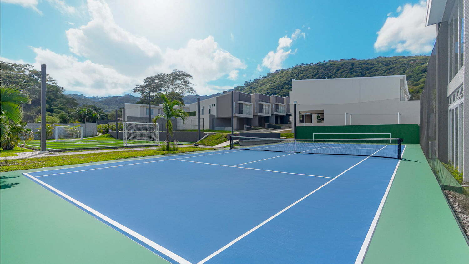 Cancha de tenis azul y verde en primer plano con una cancha de fútbol pequeña al fondo, ubicadas en las amenidades de un condominio moderno rodeado de naturaleza.