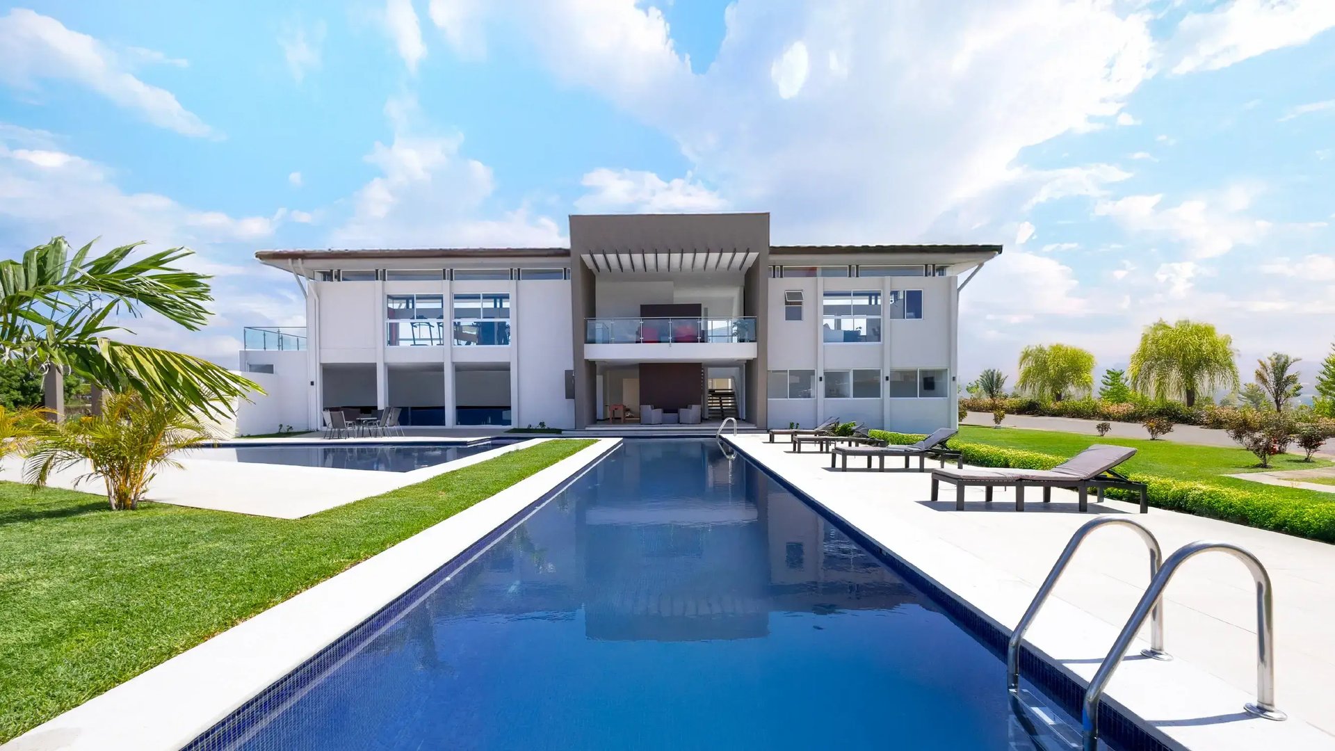 Render de la casa club de Condominio Hacienda El Cafeto de Eurohogar en Belén, Heredia. Vista frontal con una larga piscina central reflejando el edificio moderno de dos niveles con grandes ventanales, balcón central con pérgola y zona de asoleo con camastros, rodeada de palmeras y jardines.