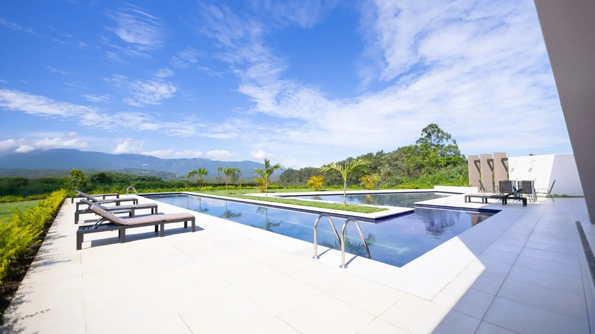 Una lujosa piscina de borde infinito en una casa club moderna con una isla central de césped y una palmera. La terraza tiene azulejos grises, tumbonas y muebles de comedor. El fondo presenta exuberantes montañas verdes bajo un cielo azul parcial.