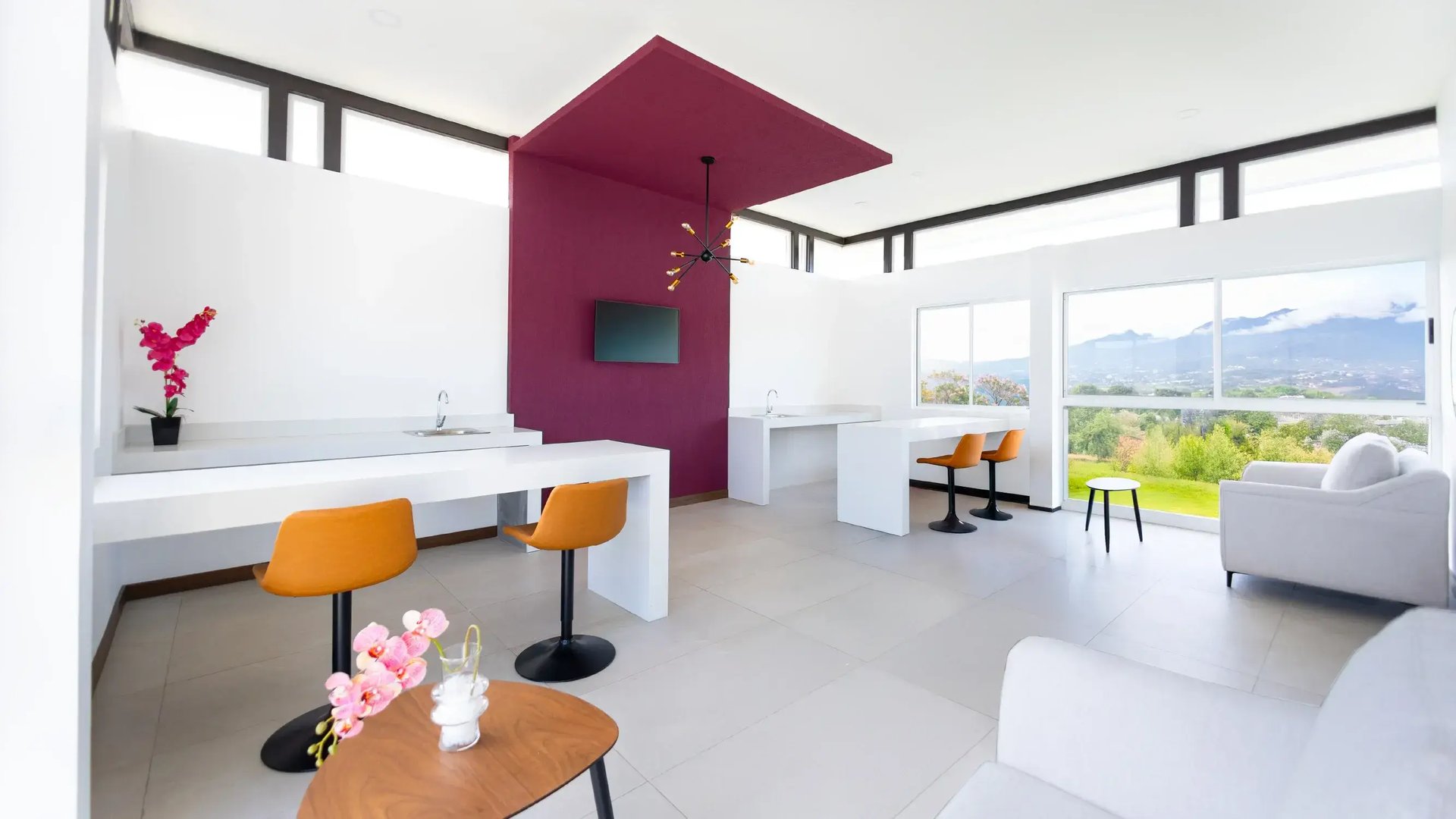 Render del área común y espacio de coworking en un condominio moderno de Eurohogar, mostrando un diseño de planta abierta con barras de trabajo blancas, taburetes color naranja, una pared central morada con TV y grandes ventanales con vista panorámica a las montañas y valles.