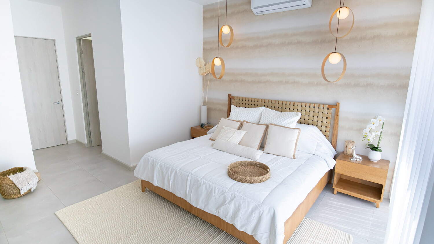 Habitación moderna en Condominio Nasara con una cama matrimonial de sábanas blancas, cabecera de madera tejida, lámparas colgantes de diseño circular y una pared decorada en tonos neutros.