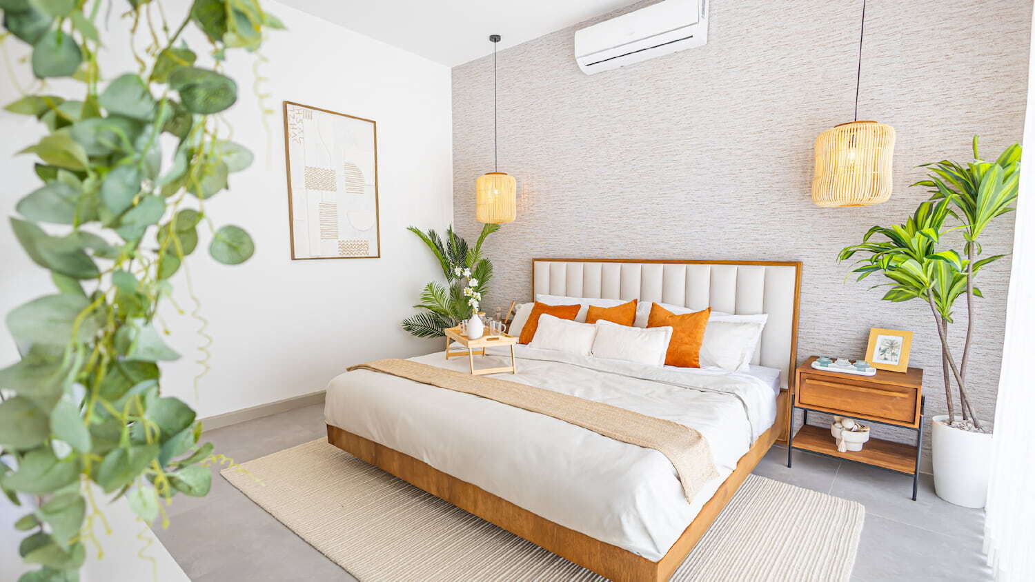 Recámara principal moderna en Condominio Nasara con una cama de sábanas blancas y cojines naranjas, lámparas colgantes de mimbre, mesa de noche de madera y plantas decorativas.