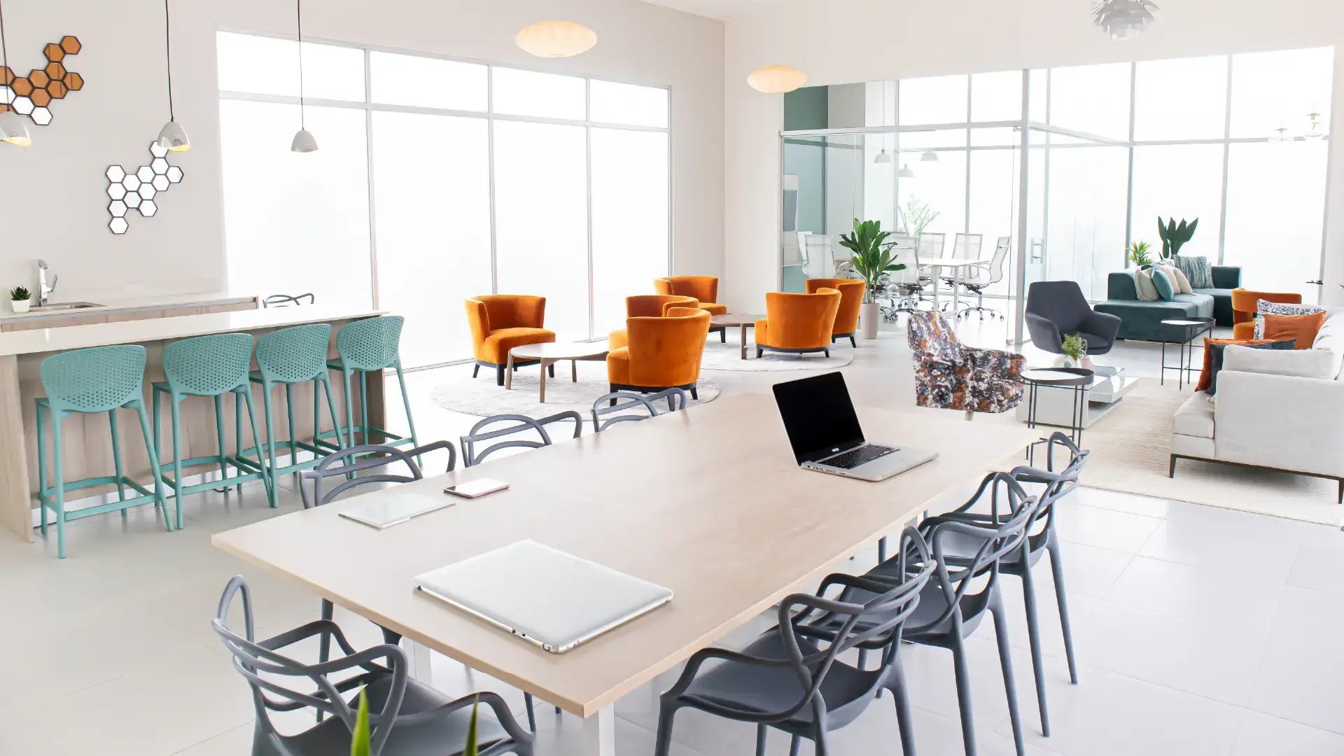 Espacio de co-working moderno en Condominio Blue, Cariari. Muestra una amplia mesa de trabajo con sillas ergonómicas, zona de lounge con sillones naranja, desayunador y salas de reuniones privadas al fondo con mucha luz natural.