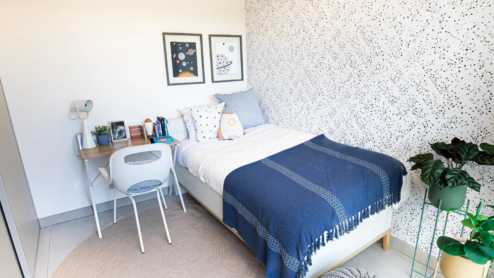 Dormitorio infantil decorado en casa modelo de Condominio Naz, Alajuela. Muestra cama individual con edredón azul, escritorio con silla blanca, papel tapiz de terrazo y cuadros temáticos espaciales.