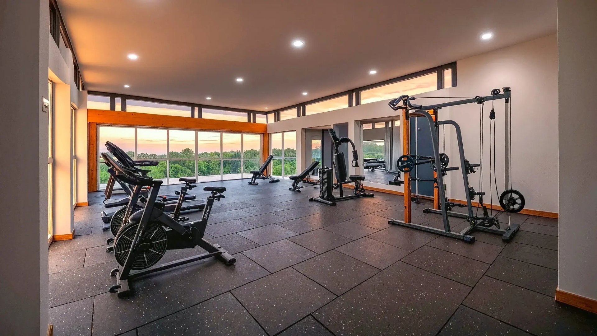 Render de un gimnasio residencial moderno y bien equipado en un condominio de Eurohogar al atardecer. Cuenta con máquinas de cardio, equipo de fuerza, piso de hule negro y grandes ventanales con vista panorámica a un paisaje boscoso.