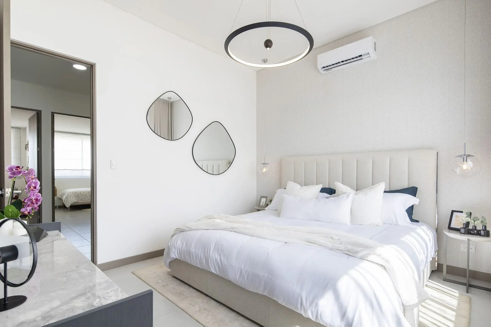 Dormitorio principal en Condominio Blue, Cariari. Muestra una cama matrimonial con cabecera acolchada, iluminación colgante de diseño, aire acondicionado, espejos decorativos y vista hacia las habitaciones secundarias. Ambiente amplio y luminoso.