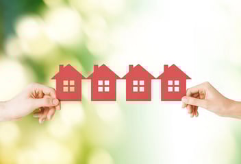 Manos sosteniendo figuras de casas rojas alineadas representando viviendas o propiedades