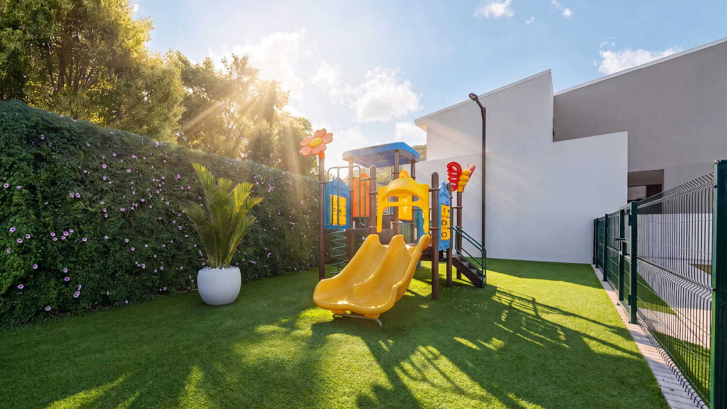 Área de juegos infantiles o playground en Condominio Nasara con un tobogán amarillo, césped sintético y rodeado por un seto verde bajo la luz del sol.