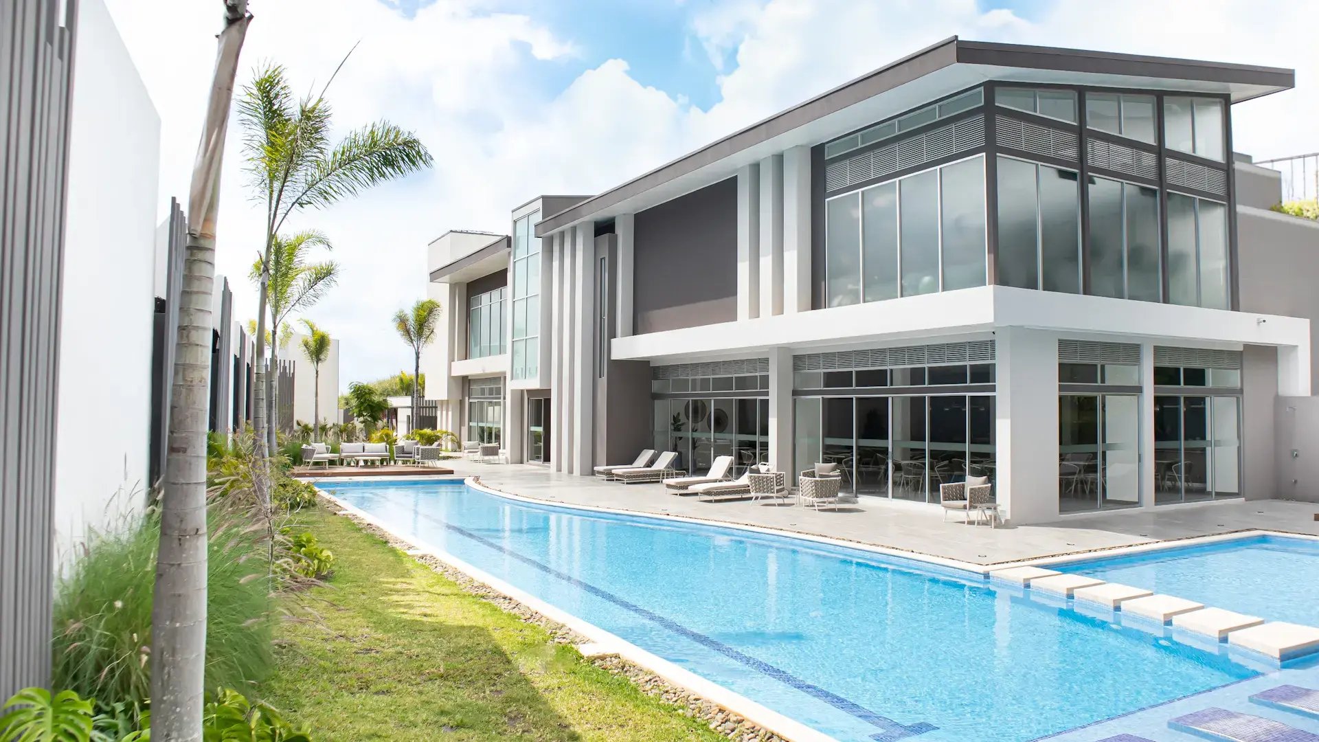 Vista exterior de la piscina tipo lap pool y la moderna casa club de dos niveles en Condominio Blue, Cariari. Se observan áreas de solárium con camastros, palmeras y arquitectura contemporánea con amplios ventanales.