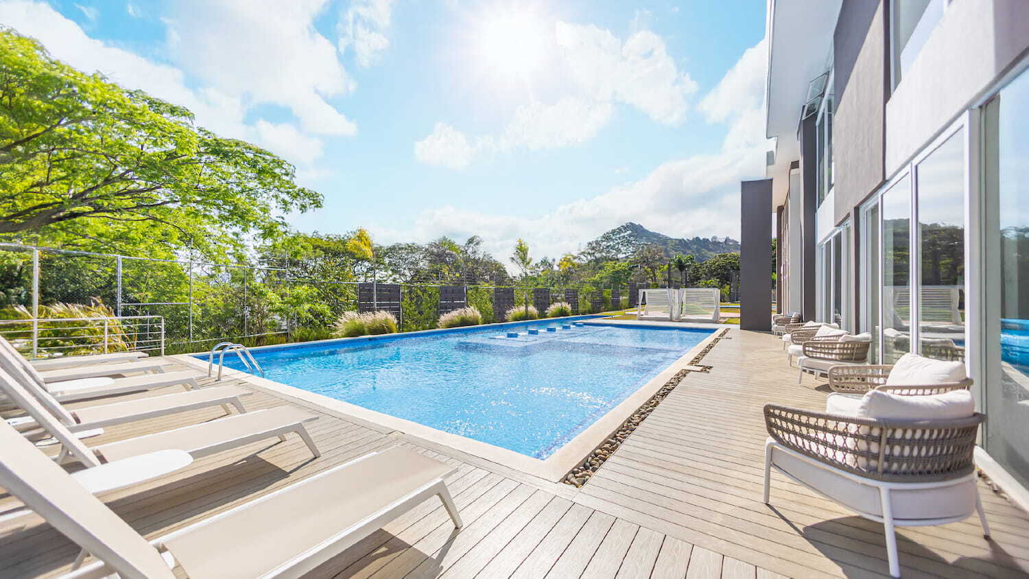 Área de piscina soleada con camastros alineados sobre una terraza de madera, sillones modernos junto al edificio y árboles bajo un cielo despejado.
