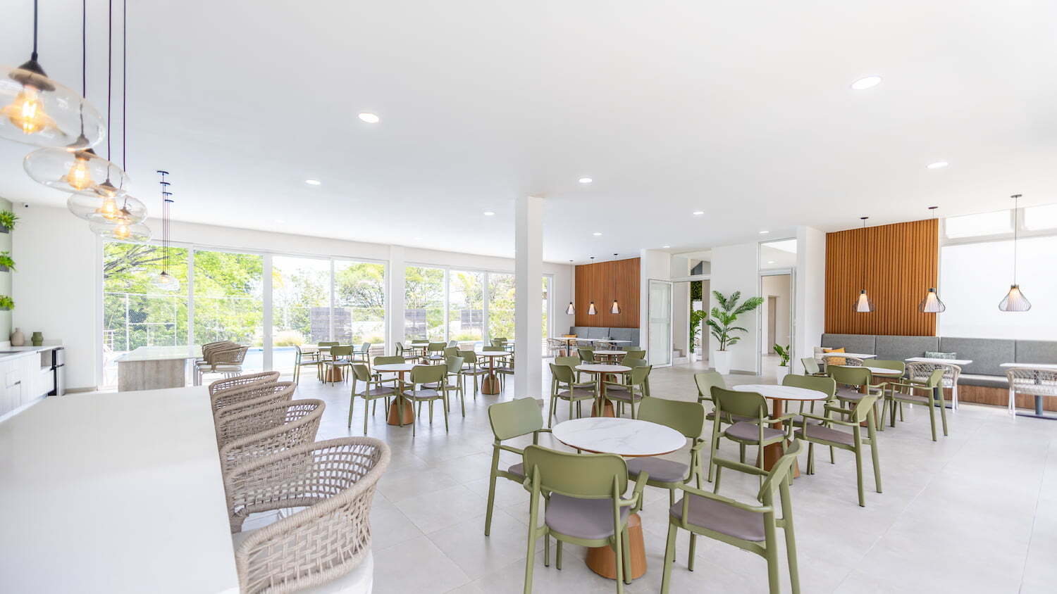 Amplio restaurante o cafetería en la casa club de Condominio Nasara con mesas redondas, sillas en tonos verdes, una barra con bancos de mimbre y grandes ventanales que iluminan el espacio.