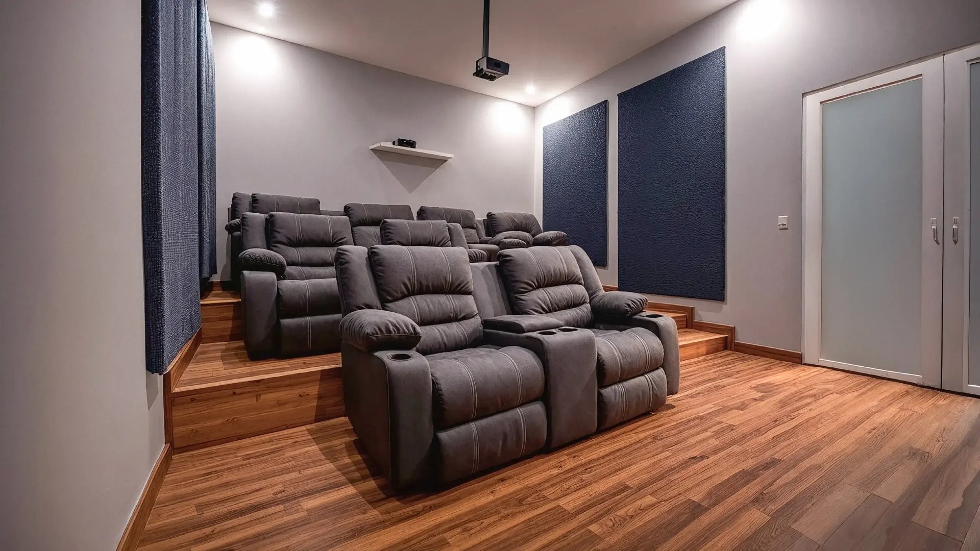 Render de una moderna y cómoda sala de cine en casa. Muestra dos filas de asientos reclinables de cuero gris con portavasos, dispuestos en un piso de madera a desnivel.