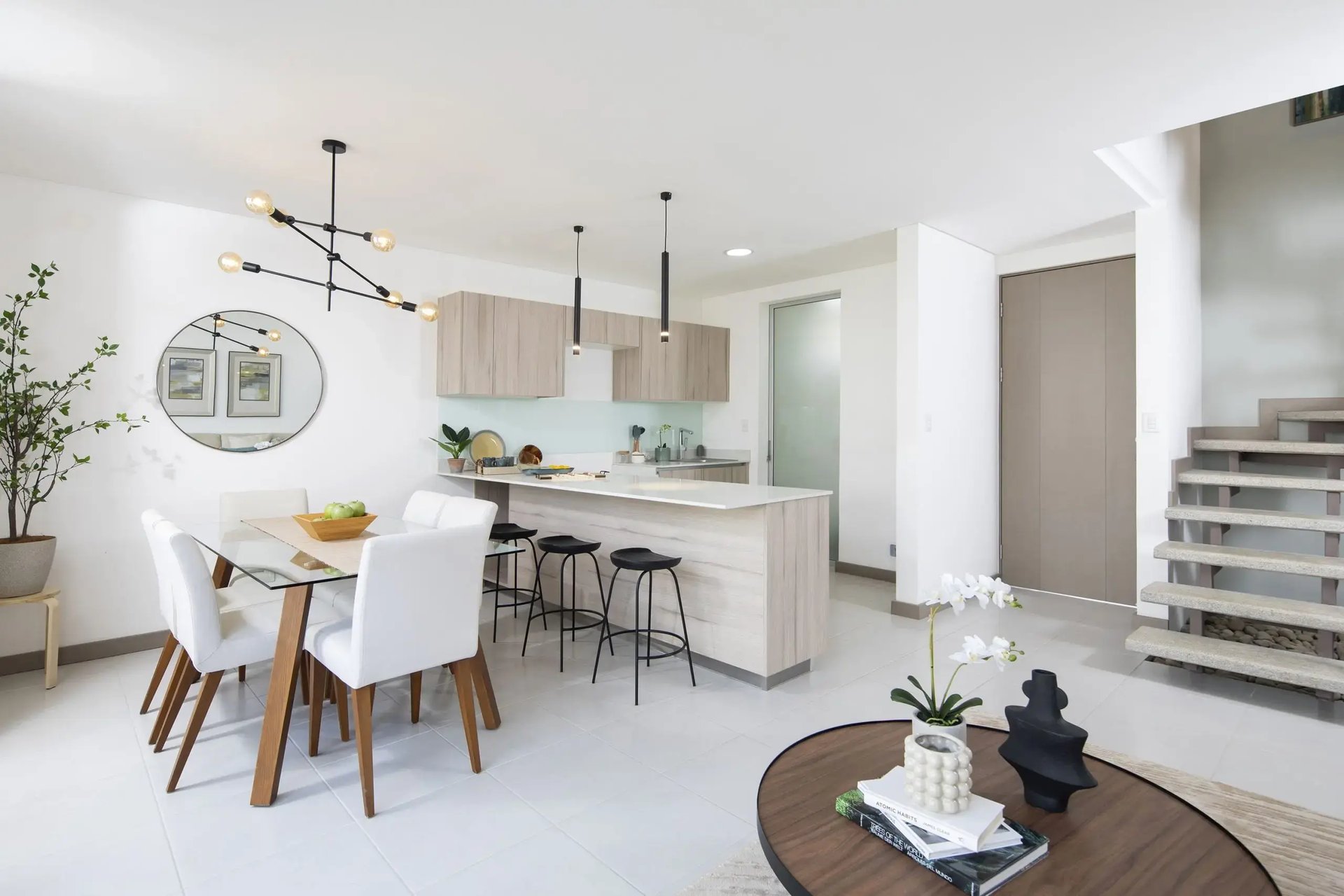 Vista interior de una casa en Condominio Blue, Cariari. Muestra el diseño de concepto abierto que integra la cocina con desayunador, el comedor con mesa de vidrio y la sala. Destaca la escalera moderna, iluminación contemporánea y acabados en tonos claros.