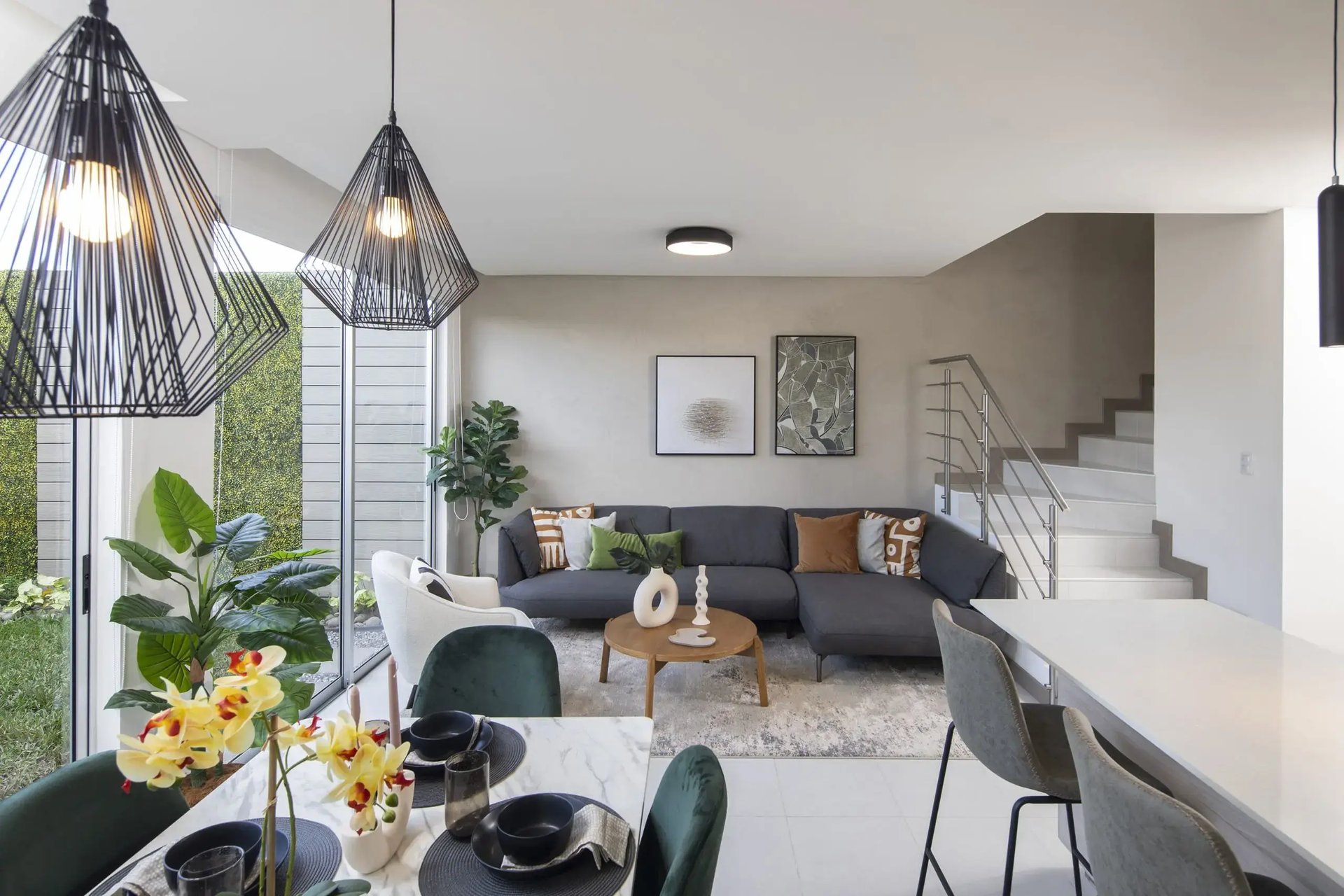 Vista panorámica de la sala y comedor en Condominio Blue, Cariari. Muestra un sofá seccional gris, lámparas de diseño industrial, comedor elegante y amplios ventanales con vista a jardines internos. Espacios modernos y funcionales de Eurohogar.