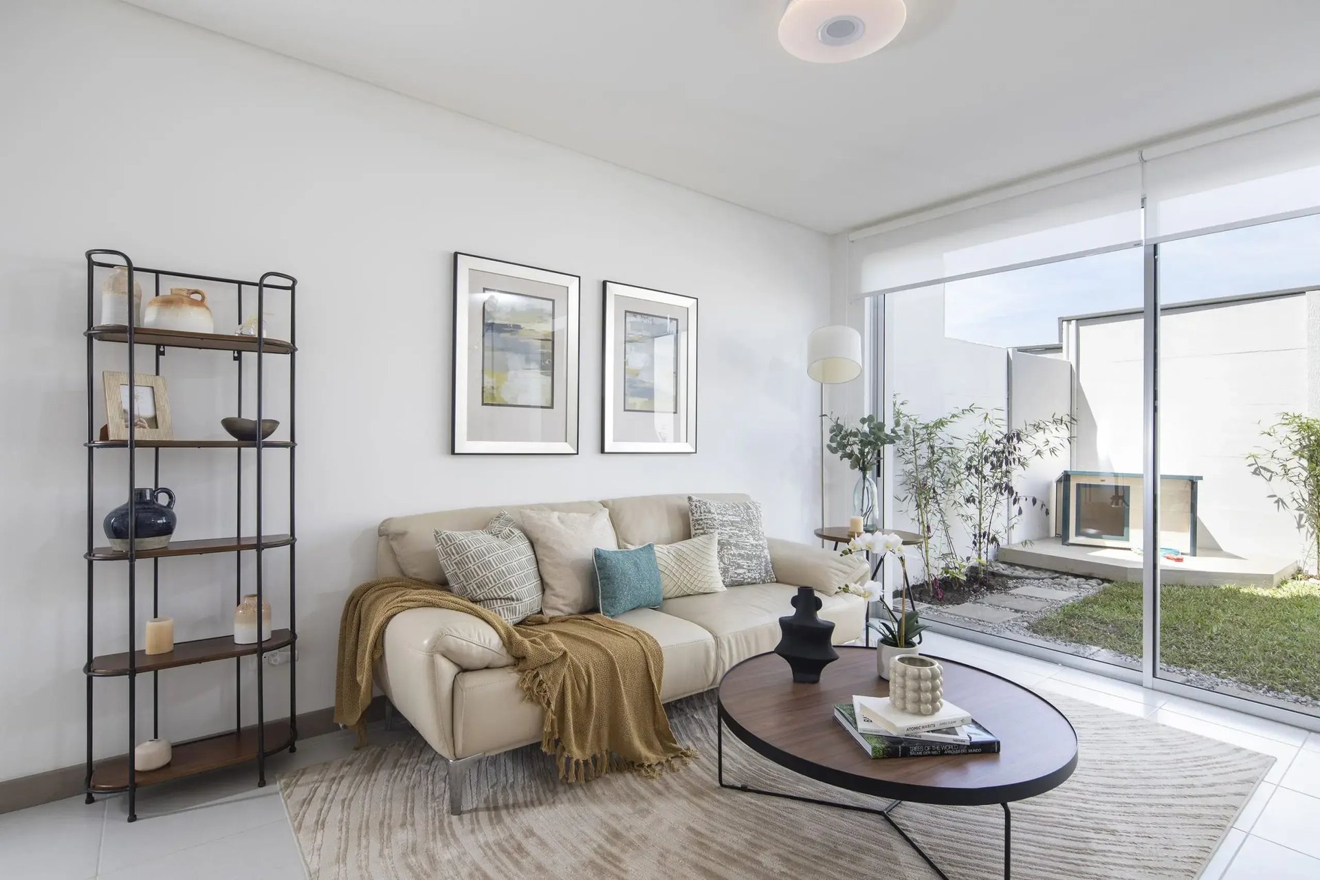 Interior de la sala en Condominio Blue, Cariari. Muestra un sofá contemporáneo en tonos crema, decoración minimalista y amplios ventanales corredizos que dan acceso a un patio privado con jardín. Espacios iluminados y modernos.