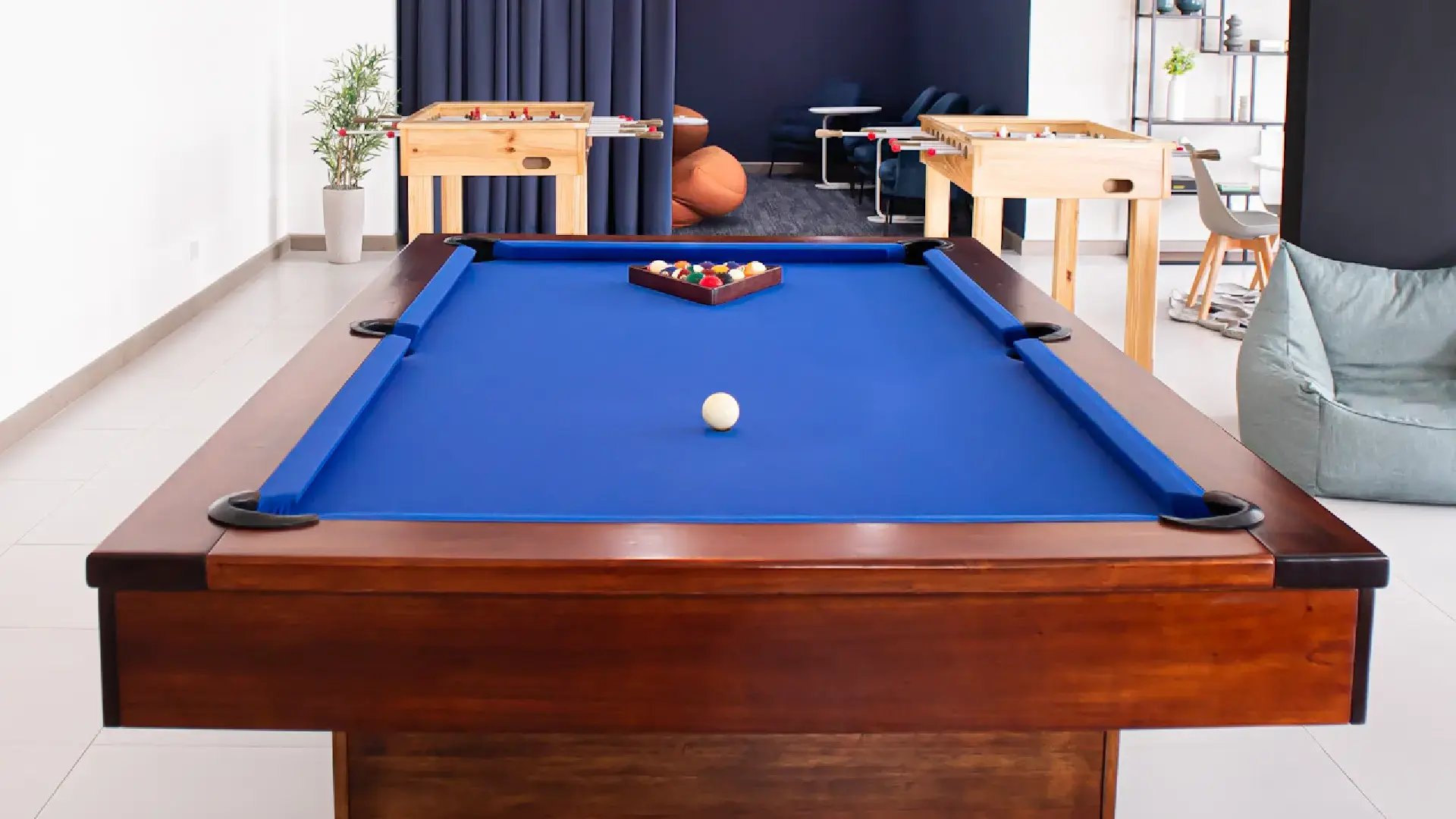 Área social y de juegos en Condominio Blue. Muestra una mesa de villar profesional con paño azul en primer plano, mesas de futbolín al fondo, y una zona de lounge con puffs y butacas. Ambiente moderno para el entretenimiento en Cariari.