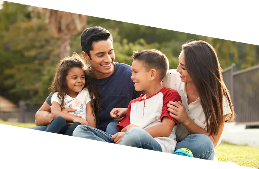 Una familia de cuatro integrantes —padre, madre y dos hijos— sentados en el césped de un parque, sonriendo y mirándose con cariño durante un día soleado.