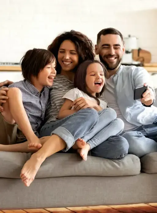Una familia de cuatro personas —madre, padre y dos hijos— sentados en un sofá gris, riendo alegremente mientras se toman una selfie con un teléfono móvil en una sala de estar moderna y luminosa.