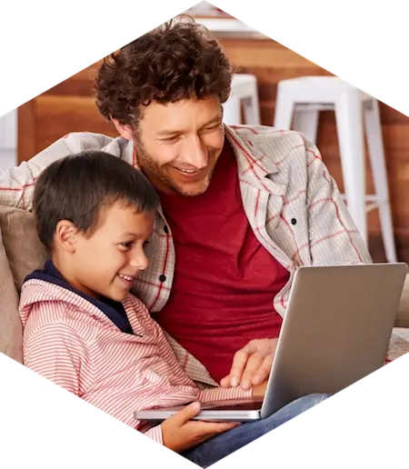 Un padre sonriente abraza a su hijo mientras ambos miran juntos la pantalla de una computadora portátil en el sofá, dentro de un encuadre hexagonal blanco.